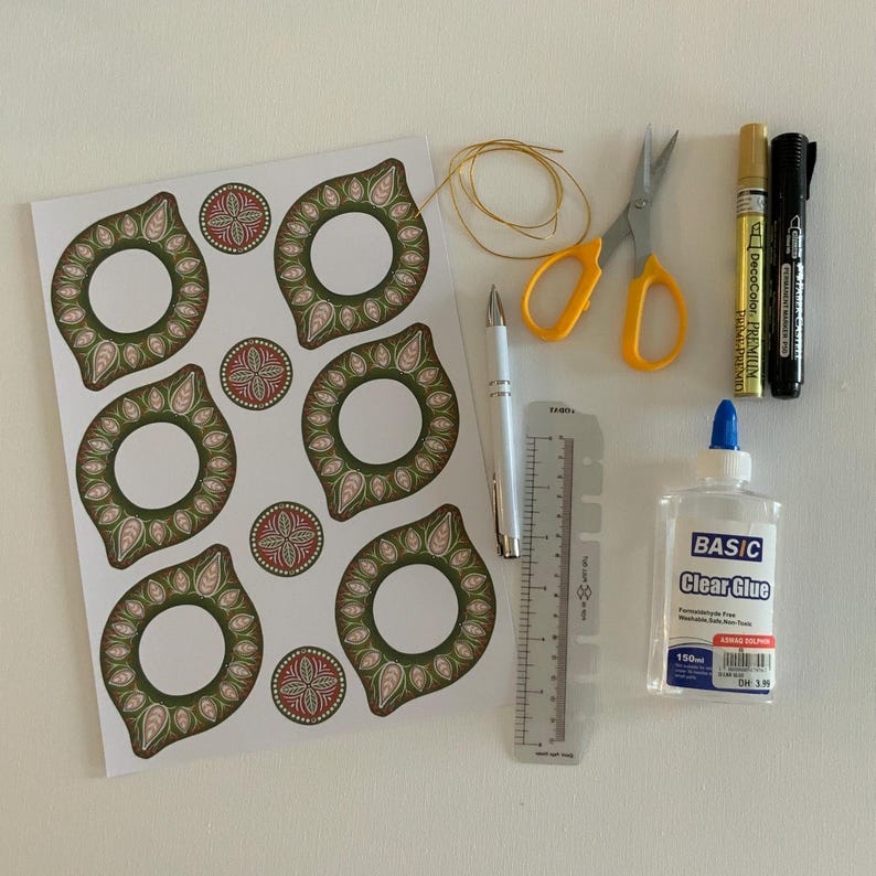 May include: An assortment of craft supplies including a sheet of decorative stickers with green and pink floral designs, scissors, pens, a ruler, clear glue, and gold string. The items are arranged on a white surface.
