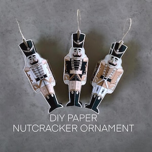 May include: Three DIY paper nutcracker ornaments. Each ornament has a detailed design with a black hat, a white moustache, and a uniform in black, white, and tan. Silver string for hanging.