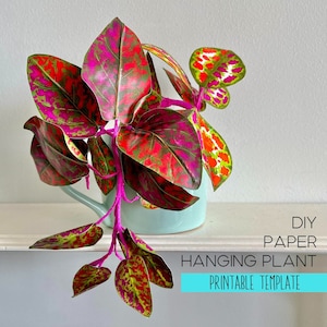 Könnte beinhalten: Eine DIY-Hängepflanze aus Papier mit leuchtend pinken, roten und grünen gemusterten Blättern an pinken Stielen, in einem hellblauen Keramiktopf. Der Text DIY PAPER HANGING PLANT PRINTABLE TEMPLATE ist unten rechts zu sehen.