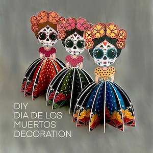 May include: Three standing paper figures featuring sugar skull faces with floral headpieces. Each figure wears a patterned bodice and a wide, pleated skirt in red, green, or blue. The text DIY DIA DE LOS MUERTOS DECORATION is printed in white.