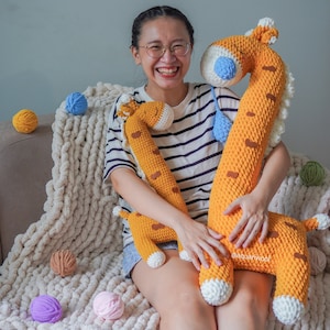 Puede incluir: Una mujer sostiene dos peluches de jirafa de crochet. La jirafa más grande es naranja y blanca con la nariz azul. La jirafa más pequeña es naranja y blanca. La mujer sonríe y está sentada en una manta blanca y esponjosa.