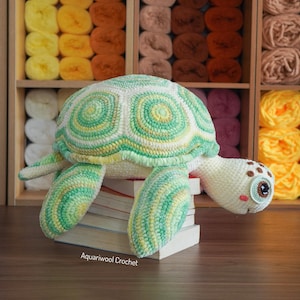 Puede incluir: Una tortuga de ganchillo hecha a mano en tonos verdes, amarillos y blancos, apoyada sobre una pila de libros. La tortuga tiene un caparazón con un patrón detallado y ojos expresivos. El fondo muestra estantes llenos de ovillos de lana.
