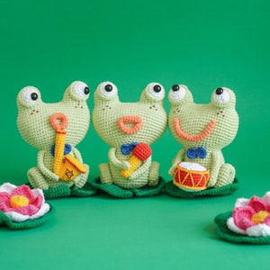 May include: Three green crocheted frog plushies playing musical instruments. The frog on the left is playing a yellow saxophone, the middle frog is playing a red tambourine, and the frog on the right is playing a red and yellow drum. Each frog is sitting on a green crocheted lily pad.