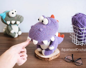 Puede incluir: Un juguete de dinosaurio de crochet morado con dientes blancos y detalles naranjas. El dinosaurio está sentado sobre una base de madera. El juguete está hecho a mano y tiene una textura moteada.