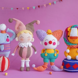 May include: Four crocheted stuffed toys: a purple and blue elephant balancing on a red and white ball, a pink and brown jester with a yellow face, a white clown with a purple and orange hat, and a yellow lion with a pink and blue mane. The lion is sitting on a blue and red platform.
