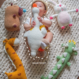 May include: A baby is surrounded by crocheted amigurumi toys, including a giraffe, a goose, a dinosaur, a bunny, and a bear. The toys are all different colors and have different facial expressions. The baby is holding the goose toy and smiling. The text "Snuggle Amigurumi" is written in the top center of the image. The text "aquariwool" is written in the bottom center of the image.