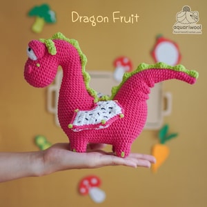 May include: A handmade, pink crochet dragon fruit toy with green detailing. The dragon fruit features a white and black design on its side, and the text "Dragon Fruit" is above it. The toy is held in a hand.