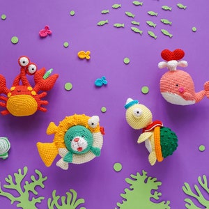 Könnte beinhalten: Ein Set aus fünf gehäkelten Meereswesen auf einem violetten Hintergrund. Die Kreaturen umfassen einen Krebs, einen Wal, eine Schildkröte und zwei Fische. Die Kreaturen sind alle bunt und haben freundliche Gesichter.