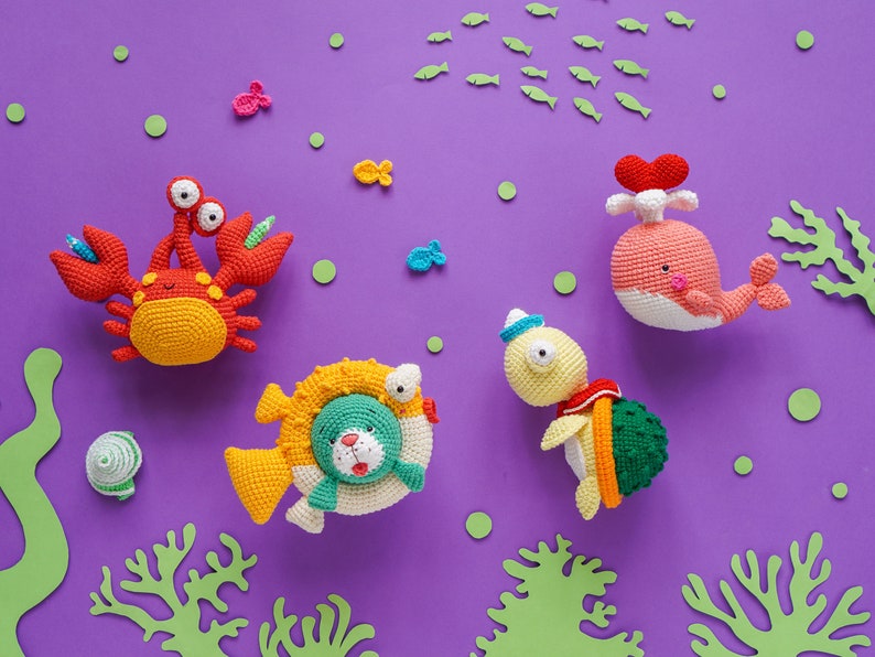 K&ouml;nnte beinhalten: Ein Set aus f&uuml;nf geh&auml;kelten Meereswesen auf einem violetten Hintergrund. Die Kreaturen umfassen einen Krebs, einen Wal, eine Schildkr&ouml;te und zwei Fische. Die Kreaturen sind alle bunt und haben freundliche Gesichter.