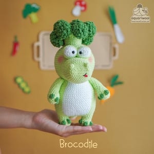 May include: A crocheted Brocodile toy, a whimsical creature with a broccoli head and a crocodile body. The toy is primarily light green with a white belly, large white eyes, and pink cheeks. The word "Brocodile" is written at the bottom.