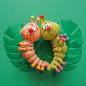 Può includere: Due giocattoli a forma di bruco all'uncinetto, uno arancione e uno verde, con facce ricamate e dettagli colorati. I giocattoli sono su uno sfondo verde con un ritaglio di carta di una foglia di monstera.