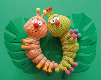 May include: Two crocheted caterpillar toys, one orange and one green, with embroidered faces and colorful details. The toys are lying on a green background with a paper cutout of a monstera leaf.