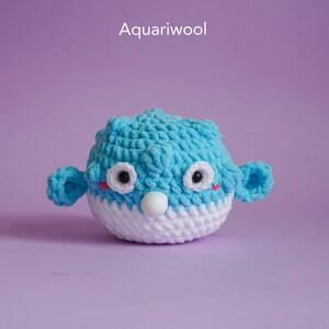 May include: A blue and white crocheted pufferfish toy with black eyes and red cheeks. The toy is sitting on a purple background.