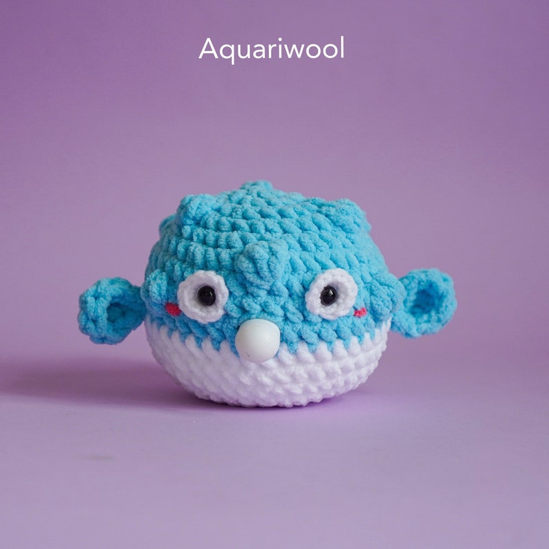 May include: A blue and white crocheted pufferfish toy with black eyes and red cheeks. The toy is sitting on a purple background.