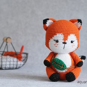 May include: A crocheted orange fox plush toy with black eyes and a green leaf in its paws. The fox is sitting on a gray surface.
