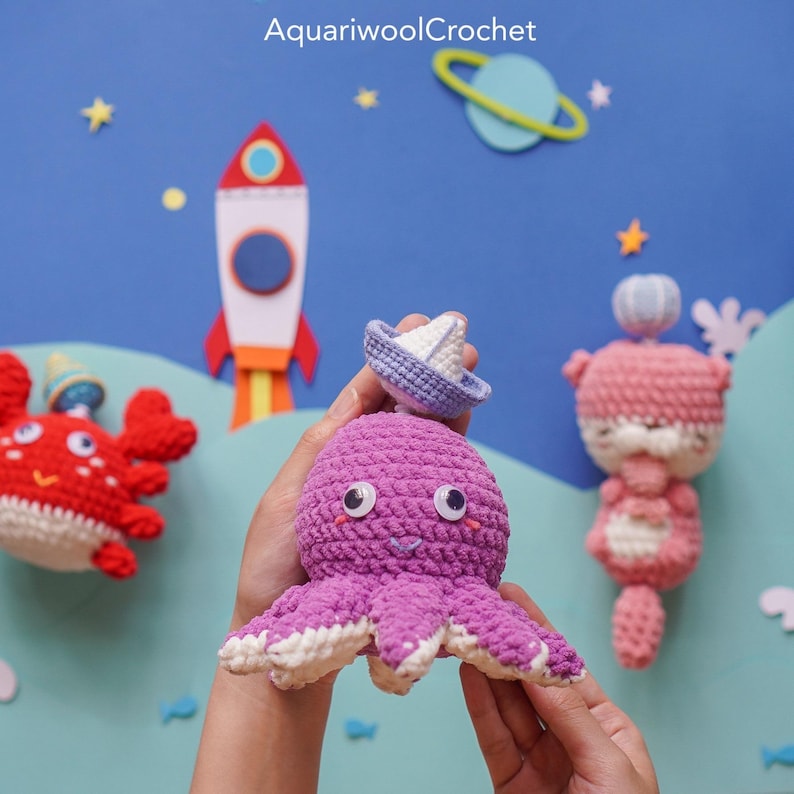 May include: A crocheted purple octopus wearing a blue and white sailboat hat. The octopus has white eyes and a smiling mouth. The octopus is being held by a person's hands against a blue background with stars and a rocket ship.