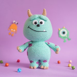 May include: A crocheted green monster toy with purple spots and large black eyes. The toy is standing on a purple surface with small colorful beads scattered around it.
