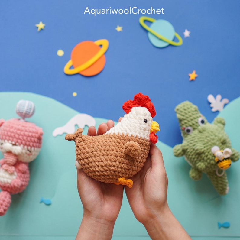 May include: A brown and white crocheted chicken toy with a red comb and yellow beak. The chicken is being held in the hands of a person. The background is blue with stars and planets.