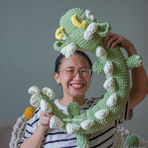 May include: A person holding a green and white crocheted plush toy shaped like a dragon. The toy has a smiling face with yellow eyes and a white mouth. The person is wearing a striped shirt and is smiling.