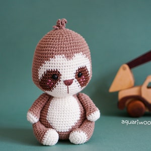 May include: A brown and white crocheted sloth toy with black eyes and pink cheeks. The sloth is sitting on a green surface with a wooden toy truck in the background.