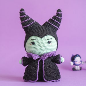 Peut inclure: Un jouet en peluche crocheté de Maléfique de La Belle au bois dormant de Disney. Le jouet est vert avec des cornes violettes et une robe noire. Le jouet a une expression espiègle sur son visage.