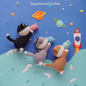 May include: Three crocheted cat toys in different colours, each holding a small crocheted fish toy. The cats are flying in a blue sky with stars, planets, and a rocket ship.