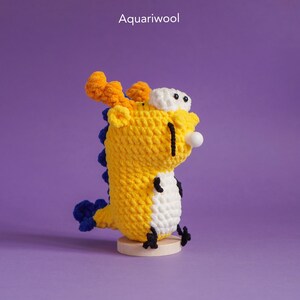 May include: A yellow crocheted dragon with orange horns, white eyes, and a blue tail. The dragon is sitting on a wooden base.