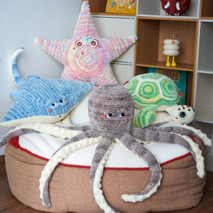 Peut inclure: Collection d'animaux marins crochetés à la main, dont une étoile de mer, une raie, une pieuvre et une tortue, disposés sur un pouf marron et blanc. L'étoile de mer est de couleur pastel, et la pieuvre est grise et blanche.