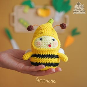 May include: A yellow and black crochet bee plush toy, with a banana-shaped hood. The toy has a smiling face with a red tongue sticking out. The word "Beenana" is written at the bottom of the image.