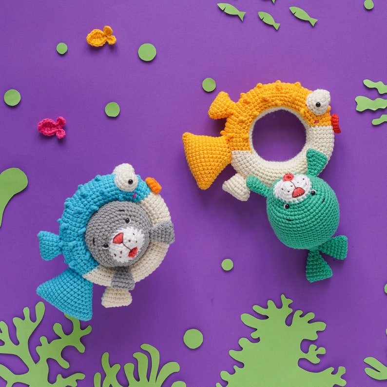 K&ouml;nnte beinhalten: Zwei geh&auml;kelte Stofftiere, ein blaues und graues Seeh&uuml;ndchen, das einen blauen und wei&szlig;en Ring h&auml;lt, und ein gr&uuml;nes Seeh&uuml;ndchen, das einen gelben und wei&szlig;en Ring h&auml;lt. Die Spielzeuge befinden sich auf einem violetten Hintergrund mit gr&uuml;nen und wei&szlig;en Papierschnitten.