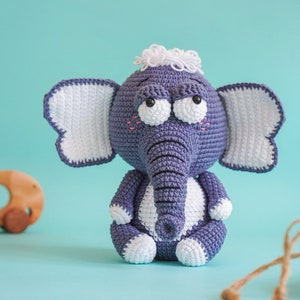 May include: A crocheted elephant toy with a grey body, white ears, and a white tuft of hair on its head. The elephant has large, black eyes and a long, grey trunk.