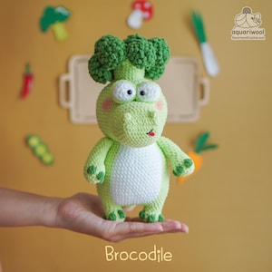 Op de afbeelding: Een gehaakt Brocodil-speeltje, een grillig wezen met een broccolihoofd en een krokodillichaam. Het speelgoed is voornamelijk lichtgroen met een witte buik, grote witte ogen en roze wangen. Het woord "Brocodile" staat onderaan.