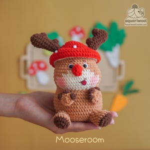 May include: A handmade crochet Moose with a red mushroom cap and brown antlers. The plush toy has a white face, pink cheeks, and a red nose and tongue. The word "Mooseroom" is written at the bottom.