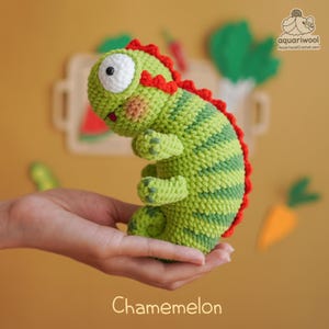 Op de afbeelding: Een handgemaakt groen en rood gehaakt kameleon speelgoed met een gestreept patroon. Het speelgoed heeft een groot wit oog, een rode rug en het woord "Chamemelon" onderaan. Het speelgoed wordt in een hand gehouden.