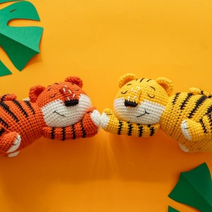 Puede incluir: Dos peluches de tigre de crochet, uno naranja y negro y otro amarillo y negro, tumbados sobre una superficie amarilla. Ambos peluches tienen los ojos cerrados y están durmiendo.