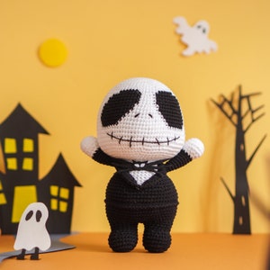 May include: A crocheted Jack Skellington doll wearing a black suit and a white bow tie. The doll is standing on a table with a yellow background.