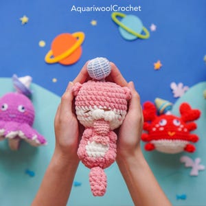 May include: A pink crocheted amigurumi otter holding a blue and white crocheted ball. The otter has white eyes and a white crocheted belly. The otter is being held by hands against a blue background with stars and planets.