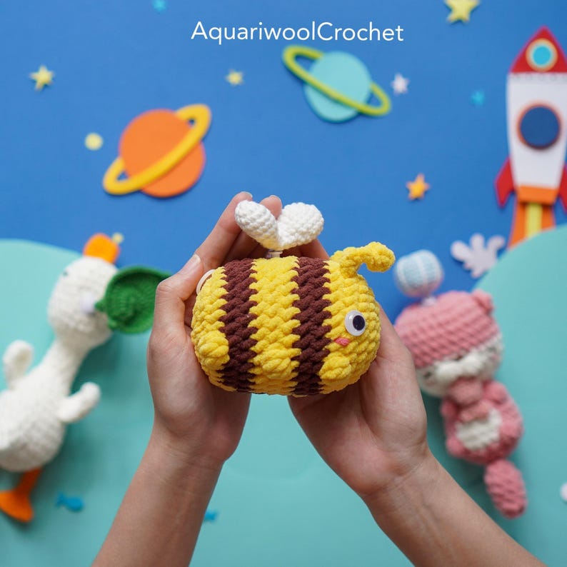 May include: A crocheted bee toy with yellow and brown stripes, white wings, and black eyes. The bee is being held in the hands of a person.