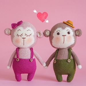 May include: Two crocheted monkey plushies holding hands. The female monkey is wearing a pink dress and a pink bow. The male monkey is wearing a brown hat and green overalls. A red heart with an arrow is above them.