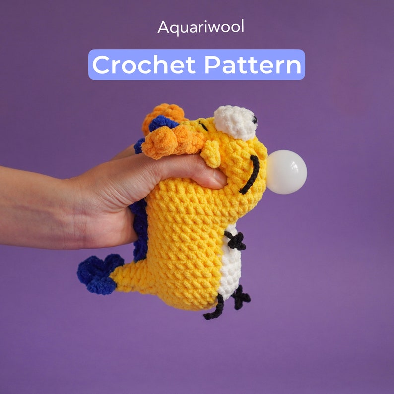 May include: A crocheted yellow and blue sea creature toy with a white face and black eyes. The toy is holding a white bubble in its mouth. The toy is being held by a hand against a purple background. The text "Aquariwool Crochet Pattern" is displayed above the toy.
