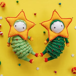 以下が含まれることがあります： 黄色い星形の帽子と緑色の体をした、かぎ針編みで作ったクリスマスツリーのオーナメントが2つ。オーナメントは黄色の背景の上に置かれています。