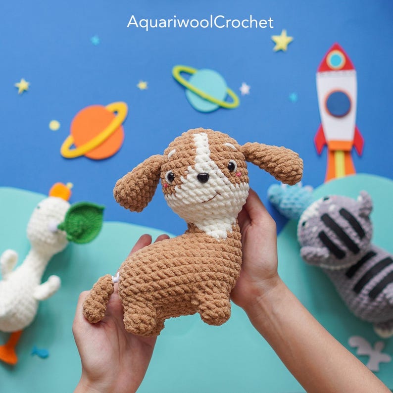 May include: A crocheted brown and white puppy toy with a white face and black eyes. The puppy is being held by a person's hands against a blue background with planets and a rocket ship.