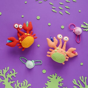 K&ouml;nnte beinhalten: Zwei geh&auml;kelte Krabbenspielzeuge mit bunten Details. Eine Krabbe ist rot und orange, die andere ist gr&uuml;n und orange. Beide haben Glitz Augen und l&auml;chelnde Gesichtsausdr&uuml;cke.