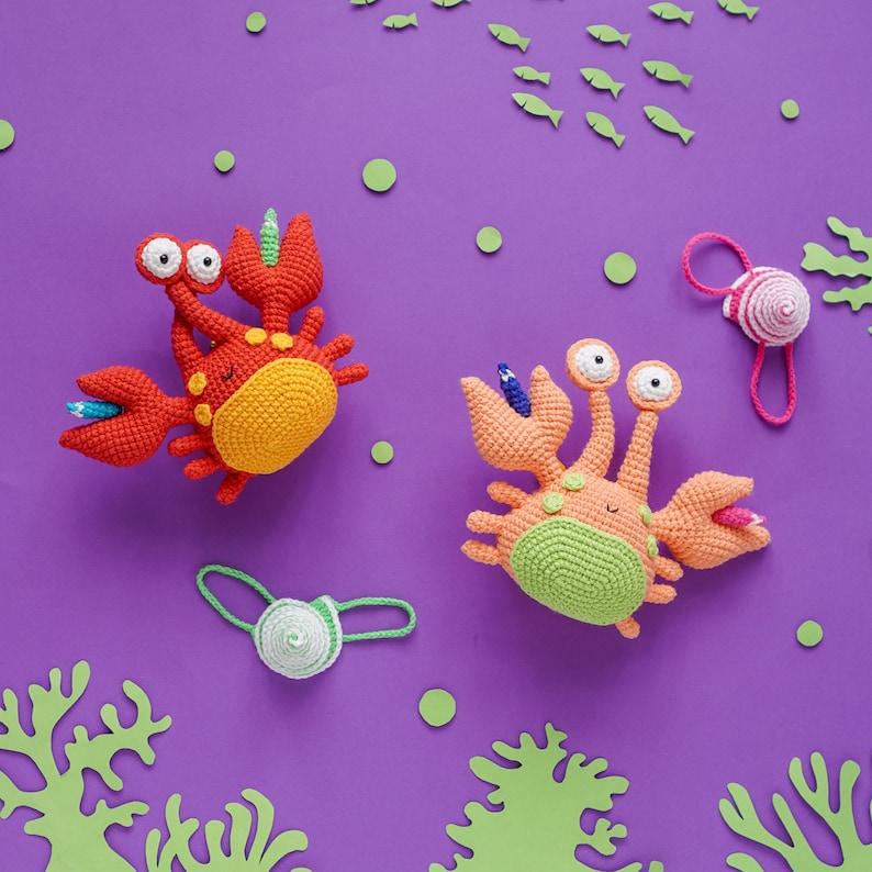 K&ouml;nnte beinhalten: Zwei geh&auml;kelte Krabbenspielzeuge mit bunten Details. Eine Krabbe ist rot und orange, die andere ist gr&uuml;n und orange. Beide haben Glitz Augen und l&auml;chelnde Gesichtsausdr&uuml;cke.