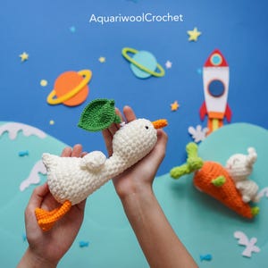 May include: A white crocheted goose with an orange beak and legs, wearing a green leaf on its head. The goose is flying in the air with a blue and green planet in the background. The text "AquariwoolCrochet" is visible in the top center of the image.
