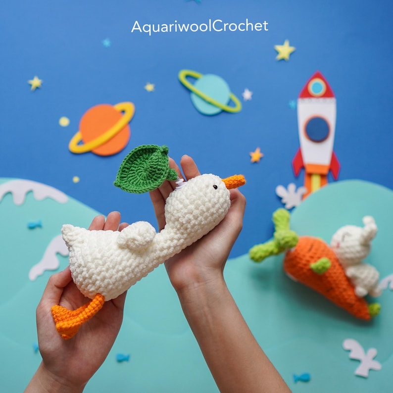 May include: A white crocheted goose with an orange beak and legs, wearing a green leaf on its head. The goose is flying in the air with a blue and green planet in the background. The text "AquariwoolCrochet" is visible in the top center of the image.