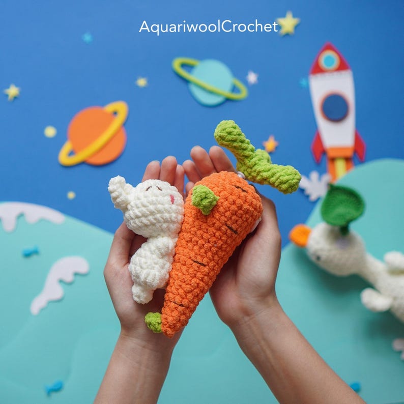May include: A pair of hands holding a crocheted carrot and a white crocheted bunny. The carrot is orange with green leaves. The bunny has a small, white nose and a pink mouth. The background is blue with white stars and planets.