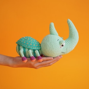 Puede incluir: Un juguete de peluche de crochet con forma de rinoceronte con cuerpo verde y patas rosas. El juguete tiene una cara amigable con un solo ojo y una pequeña boca.