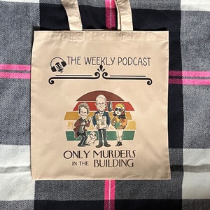 以下が含まれることがあります： レトロなデザインのベージュのトートバッグ。3人の漫画のキャラクターと「The Weekly Podcast Only Murders in the Building」というテキストが描かれています。