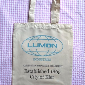 Könnte beinhalten: Eine beige Tragetasche mit einem blauen Globus-Logo, auf dem "LUMON INDUSTRIES" steht, und dem Text "MACRODATA REFINEMENT DEPARTMENT Established 1865 City of Kier".